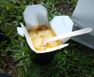 Boîte en carton remplie de truffade (fromage fondu et pommes de terres), posée sur de l'herbe.